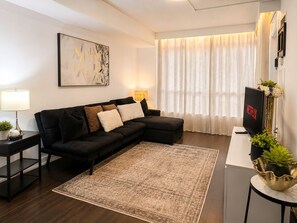 A modern living room with a sectional sofa, a flat-screen TV, a decorative rug, and a wall-mounted artwork. - The Opal Harbor Estate (Toronto)