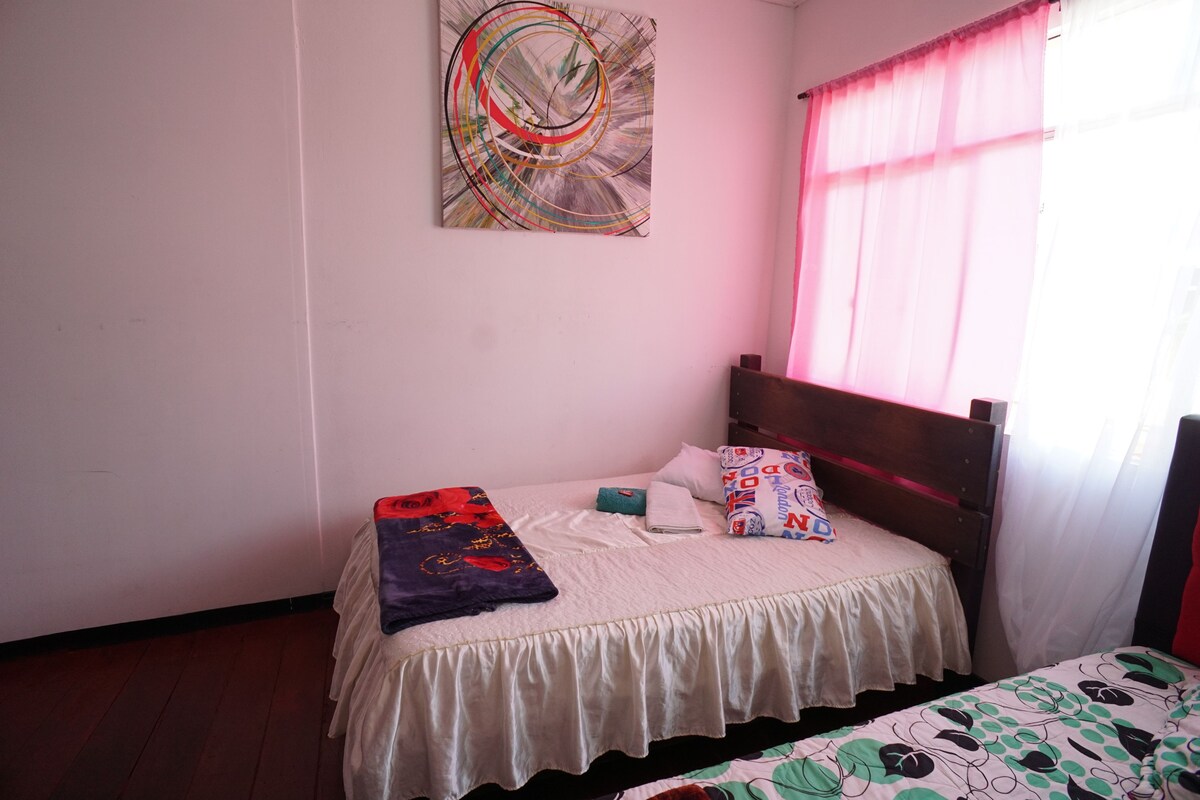 A bedroom with a single bed, a bedside table, a wall painting, and a window with curtains.