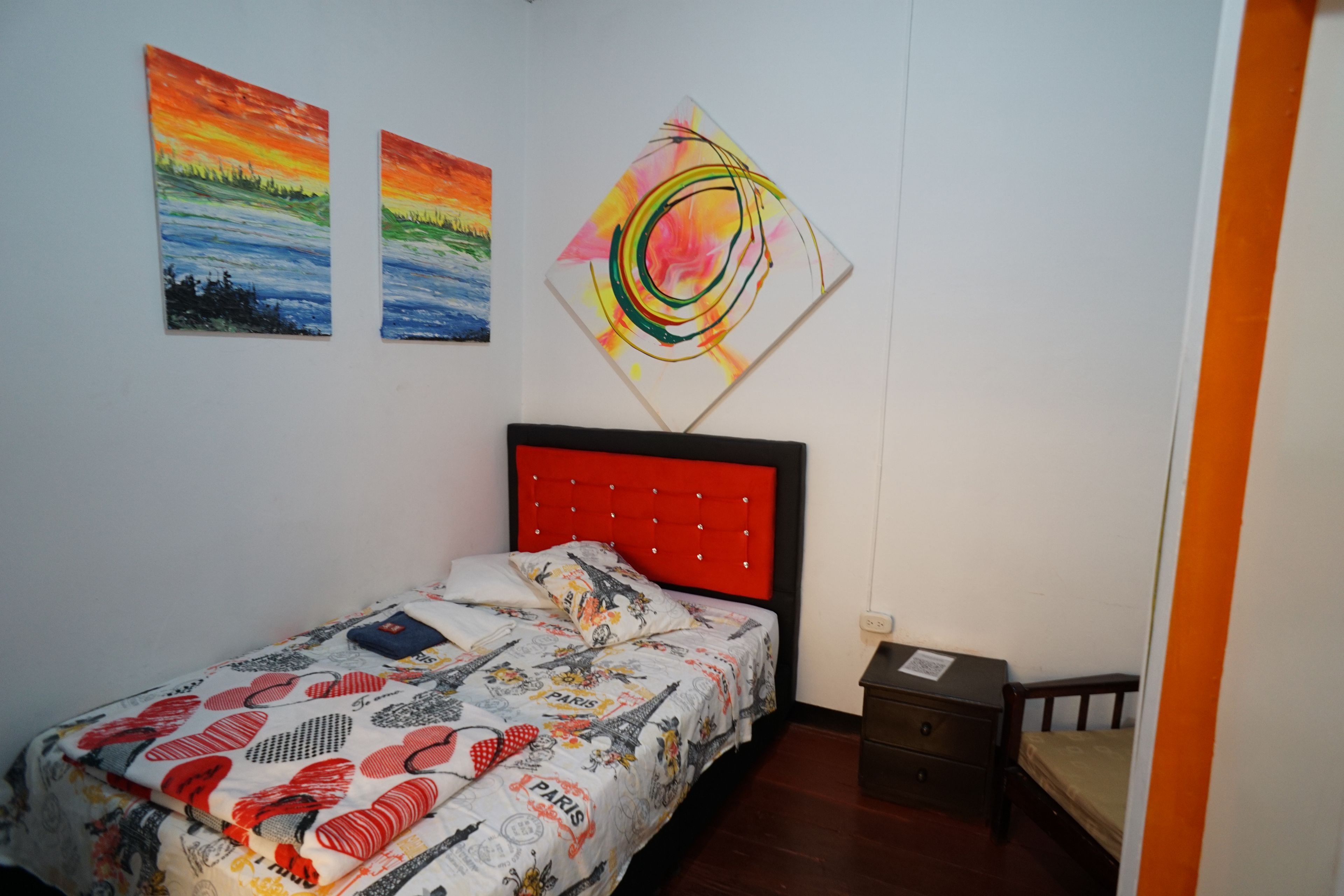 Un dormitorio con cama, mesita de noche, cajón y dos cuadros en la pared.