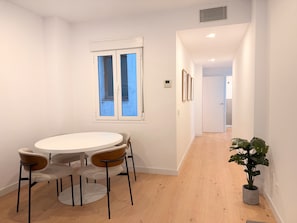 Un interior modern amb una taula rodona, quatre caires, una finestra amb persianes, una planta en test i un passadís que condueix a una altra habitació.