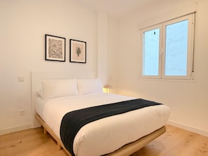 A bedroom with a bed, two framed pictures on the wall, a window, and a nightstand with a lamp. - Monteleón Apartments by Wynwood (Madrid)