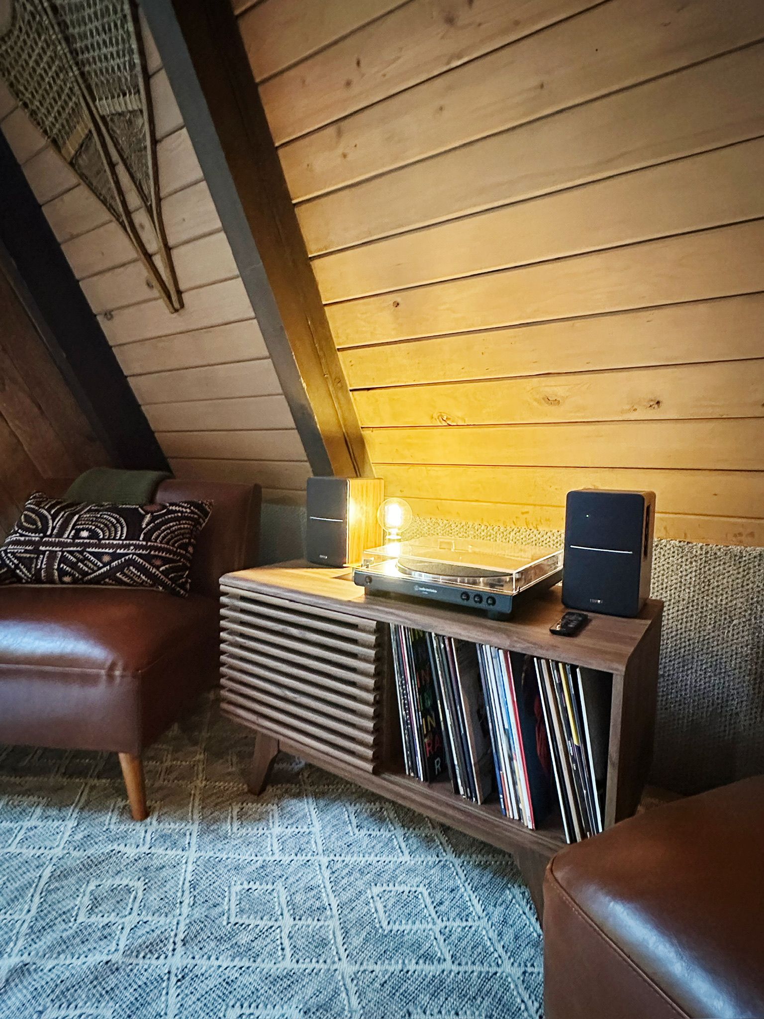 Een gezellige kamer met een houten plafond, een leren bank, een tafeltje met een lamp en een boekenkast met vinylplaten.