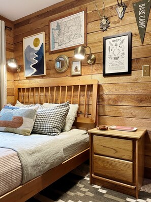 Ein Zimmer mit Holzboden, ausgestattet mit einem Bett, einem Nachttisch und Wanddekoration, darunter eine Karte und ein gerahmtes Pflanzenbild.
