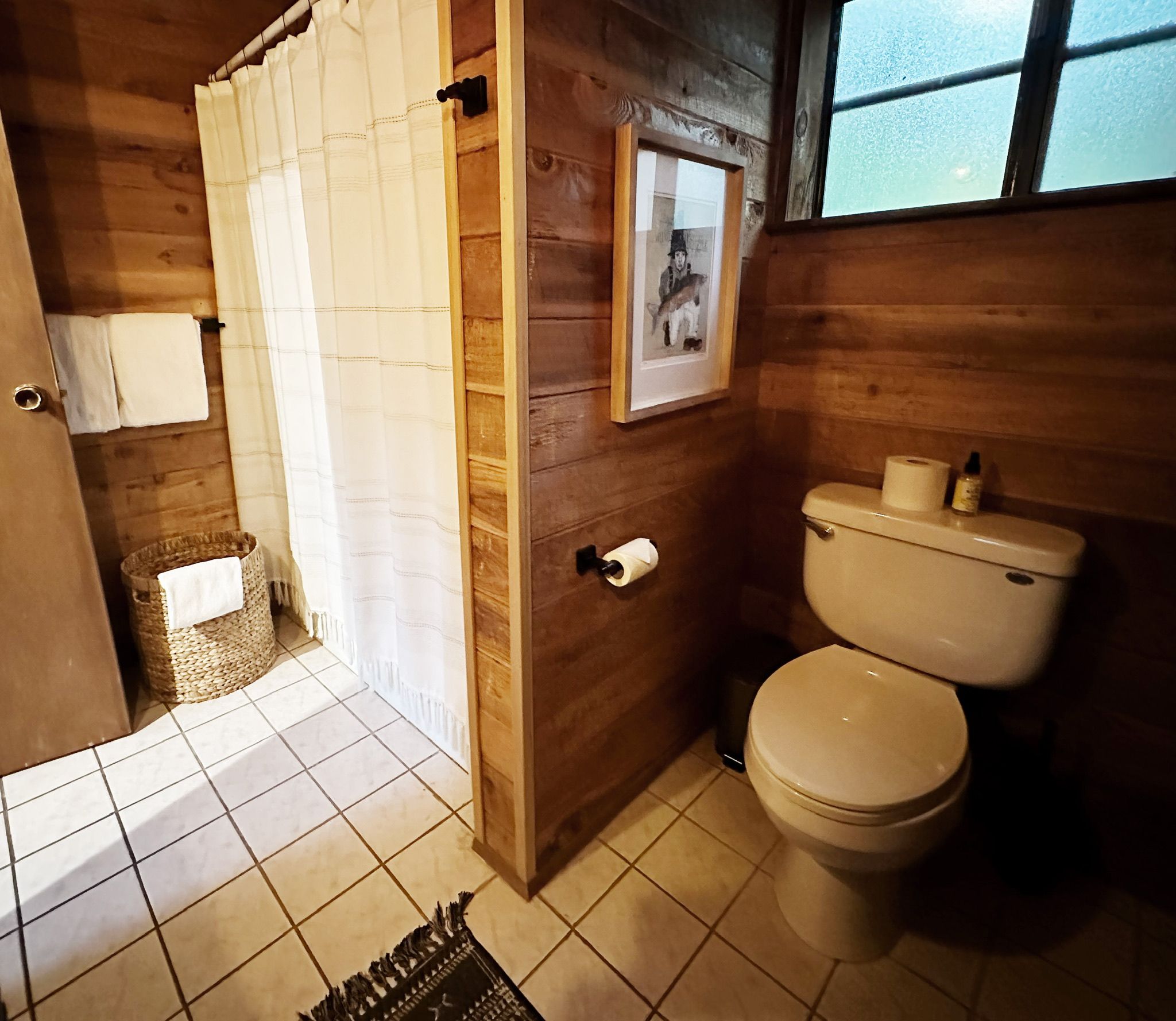Ein Badezimmer aus Holz mit Toilette, einer Duschkabine, einem gerahmten Bild an der Wand und einem Fenster.