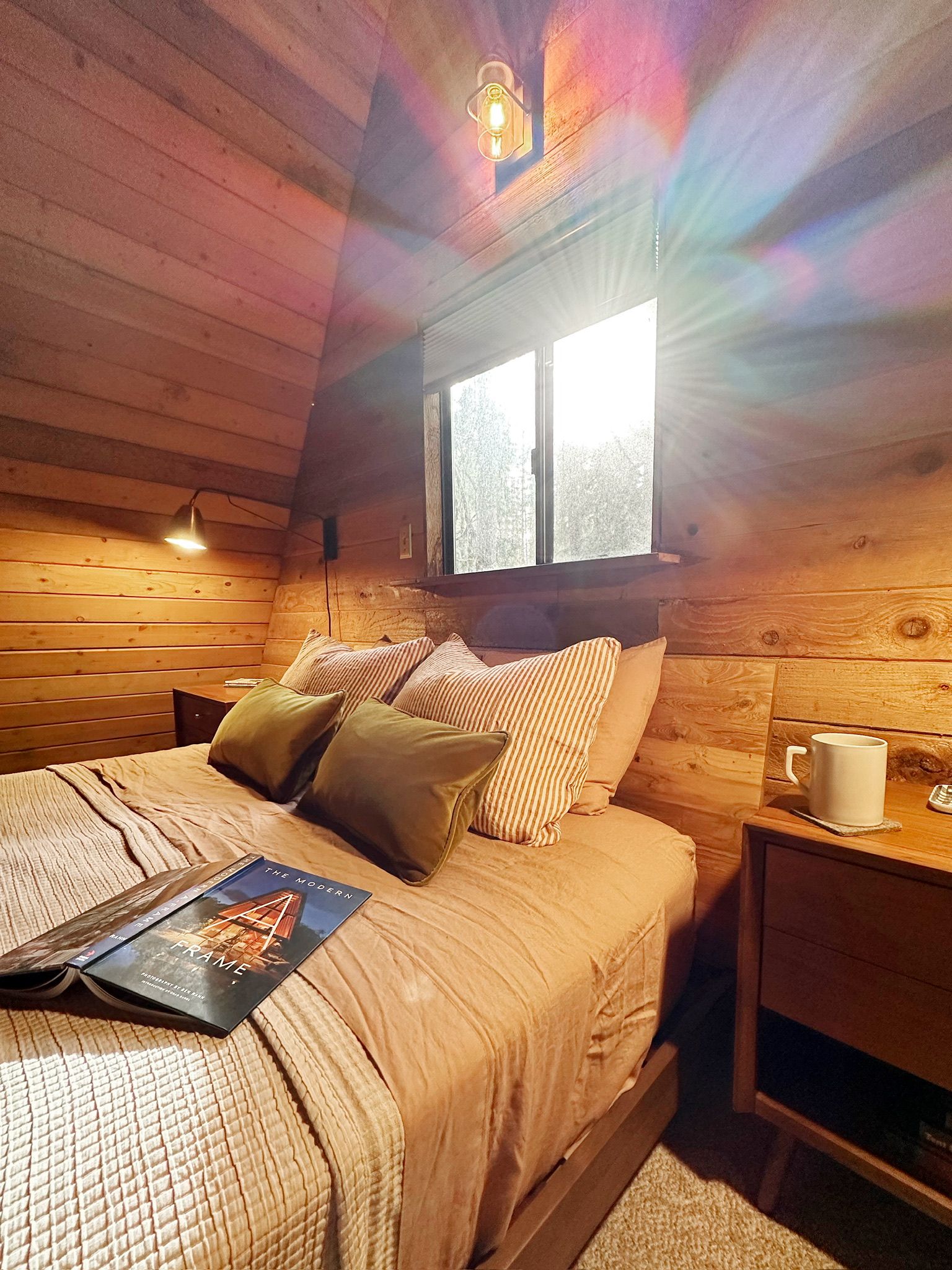 Ein gemütliches Schlafzimmer in einer Holzhütte mit einem Bett, Kissen, einem Nachttisch mit einer Tasse und einem Buch.
