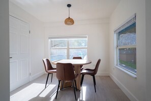 Een eethoek met een ronde tafel en zes stoelen, een groot raam met uitzicht op een achtertuin, en een hanglamp.