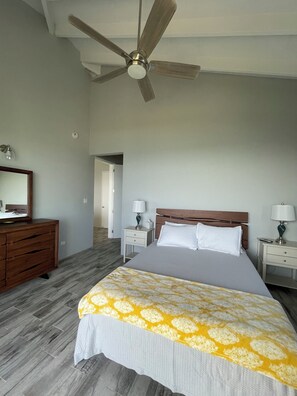 A bedroom with a bed, nightstands, a dresser, a ceiling fan, and a door leading to another room. - Modern 2BR St Croix Home with an Amazing Ocean View (Christiansted)
