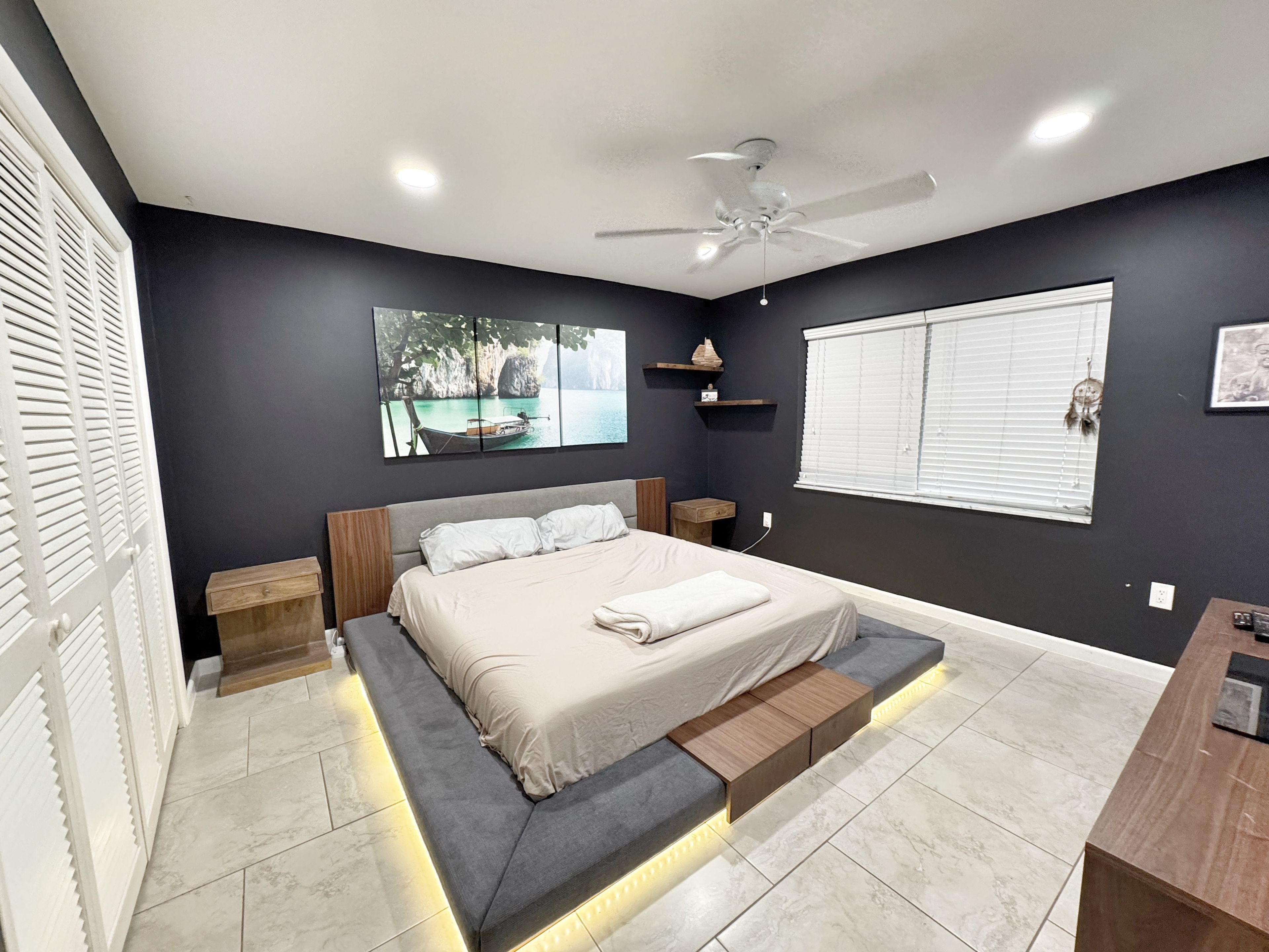 Un dormitorio moderno con cama, mesitas de noche, ventilador de techo, ventana con persianas y un estante montado en la pared con objetos decorativos.