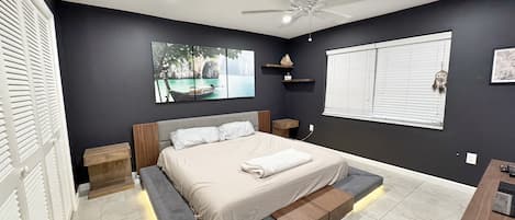 Un dormitorio moderno con cama, mesitas de noche, ventilador de techo, ventana con persianas y un estante montado en la pared con objetos decorativos.