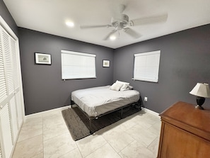 Quarto com cama, ventilador de teto e duas janelas com cortinas.