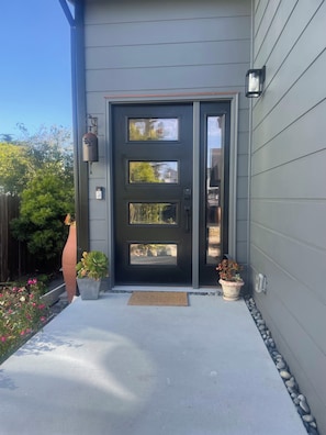 A modern house with a dark grey exterior, a black front door with three windows, a small porch with a doormat, and potted plants. - Bright + spacious home w/ sauna. Steps to ocean, bluff-top trail, and downtown (Half Moon Bay)