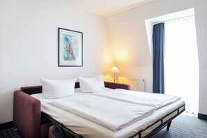 Ein Hotelzimmer mit einem Bett, einem Schreibtisch, einem Stuhl, einer Lampe, einem Fenster mit Vorhängen und einem gerahmten Bild an der Wand.