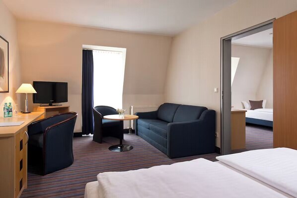 Ein Hotelzimmer mit einem Bett, einem Schreibtisch mit Lampe, einem Fernseher, einer Couch, einem kleinen Tisch und einem Fenster mit Vorhängen.
