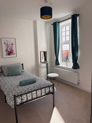 Ein Schlafzimmer mit einem Bett, einem Nachttisch, einem Sessel, einem Fenster mit Vorhängen, einer Heizung und einer Pendelleuchte.