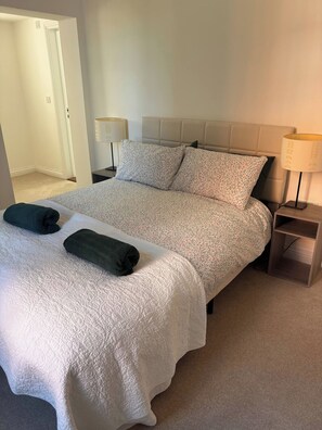 A neatly made bed with a white quilt and two dark pillows, a bedside table with a lamp, and a door leading to another room. - Charming Ground Floor Apartment Near Beach, Norfolk (Sheringham)