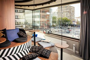 Ein modernes Hotelzimmer mit einer gebogenen Couch, einem kleinen runden Tisch und Blick auf eine belebte Straße durch große Fenster.