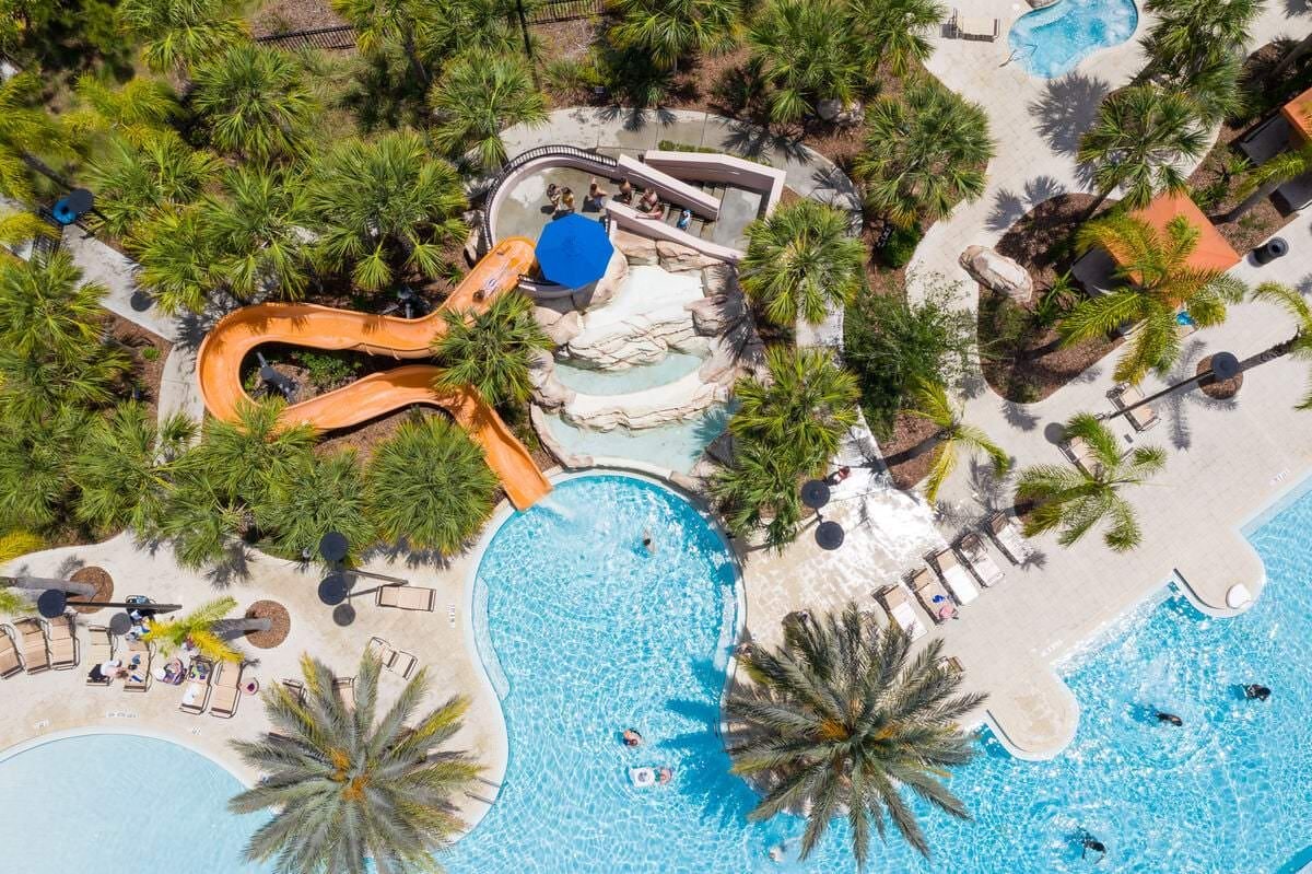 An aerial view of a resort with a large swimming pool, a water slide, and palm trees.
