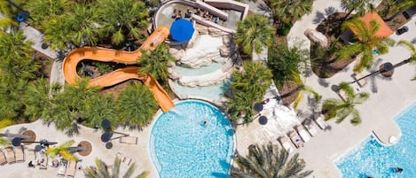An aerial view of a resort with a large swimming pool, a water slide, and palm trees.