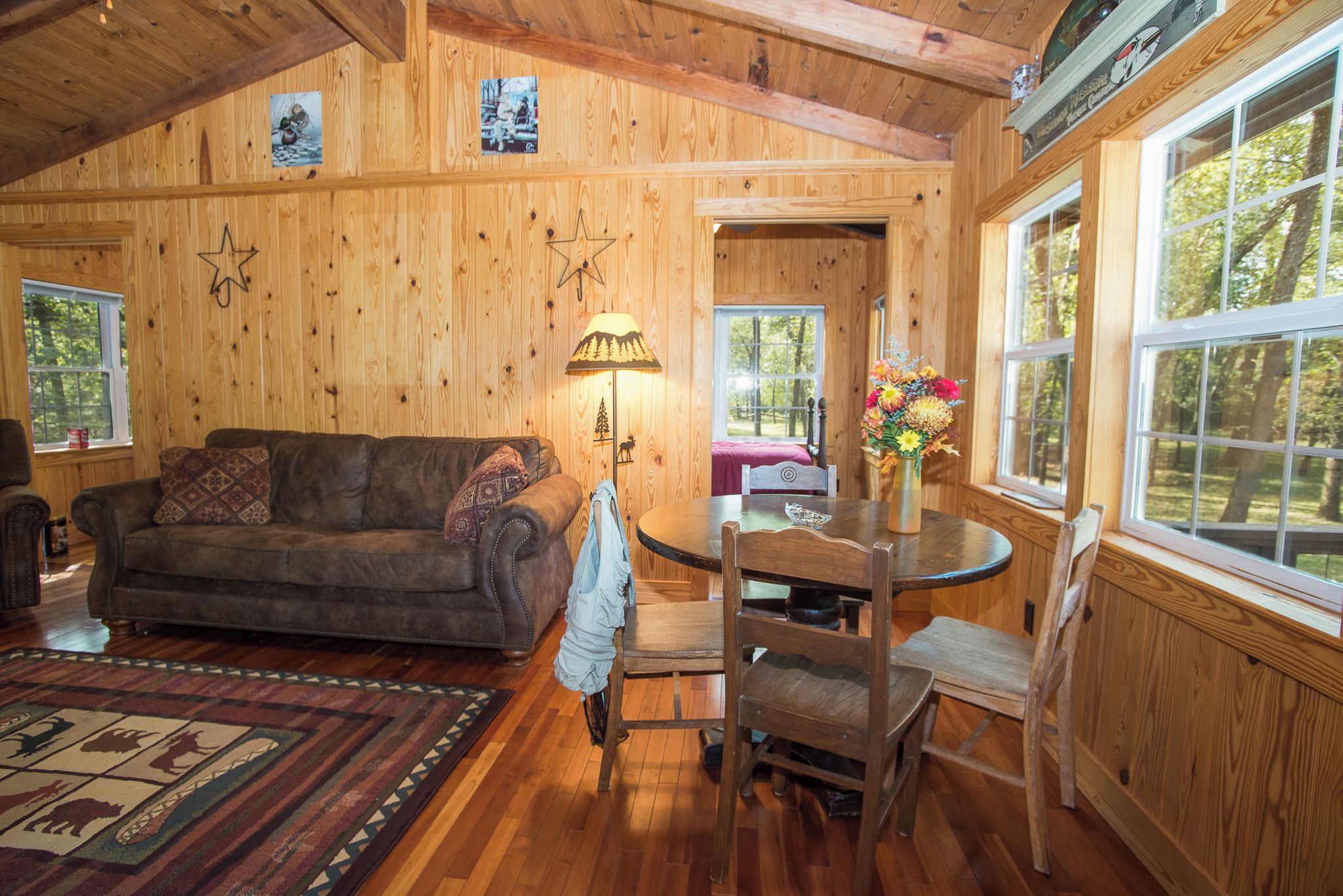 A cozy cabin interior with a wooden floor, a sofa, a dining table with chairs, and a window with a view of trees.
