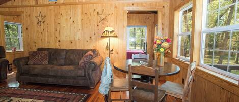 A cozy cabin interior with a wooden floor, a sofa, a dining table with chairs, and a window with a view of trees.