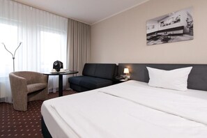 Ein Hotelzimmer mit einem Bett, einem Sessel, einem kleinen Tisch mit Wasserkocher, einer Lampe und einem Bild an der Wand.
