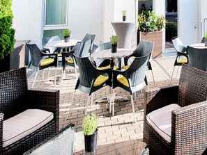 Eine Terrassengruppe im Freien mit Korbmöbeln, darunter runde Tische und Stühle, sowie Blumentöpfen.
