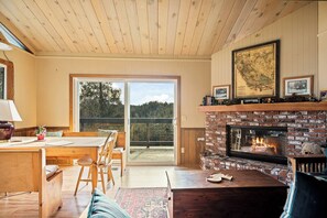 Une pièce chaleureuse avec un plafond en bois, une cheminée, un coin repas avec une table et des chaises, et une grande fenêtre donnant sur l’extérieur.