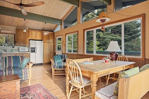 A spacious kitchen with wooden cabinets, a dining area with a wooden table and chairs, and large windows offering views of the outdoors. - Tree-Laden Crestline Cabin | 3BR + Fireplace (Crestline)
