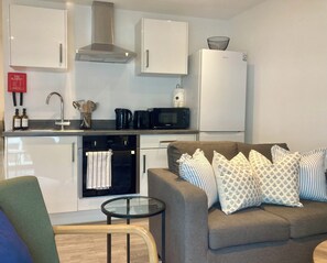 Cocina compacta con microondas, horno y gabinetes blancos. En la sala de estar, un sofá marrón con cojines estampados.