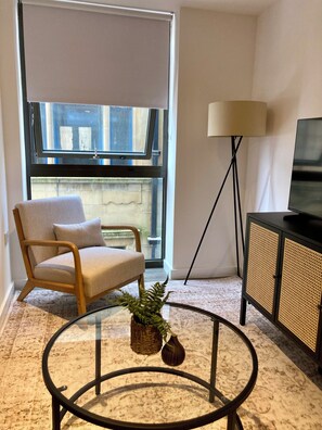 A room with a large window, a chair, a lamp, a television, and a small table with a plant. - Fargate Central 204 | 3-Bed | Sheffield Centre (Sheffield)