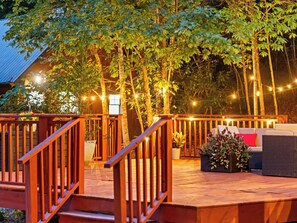 Une terrasse en bois avec une rambarde, entourée d’arbres et illuminée de guirlandes lumineuses.