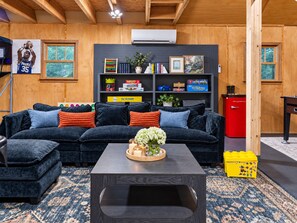 A cozy living room with a dark blue sofa, a coffee table with a floral centerpiece, and a bookshelf filled with books and games. - Private 30-Acre Retreat w/ Hot Tub, Game House/Pool Table, Trails & Pet Friendly (Old Fort)