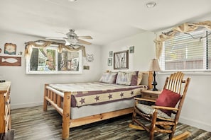 Une chambre à coucher comprenant un lit, une chaise, un ventilateur de plafond, une fenêtre avec des rideaux et des éléments de décoration murale.