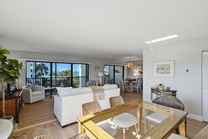 A spacious living area with a white sofa, a glass coffee table, and wicker chairs. There is a balcony with a view of palm trees and the ocean. - Sea Oats 231 | Breathe in Gulf Breezes, Unwind in Coastal Comfort & Savor Island Serenity (Boca Grande)