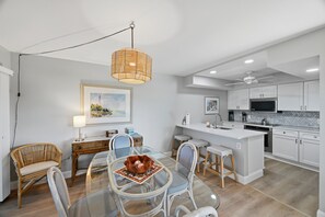 A modern kitchen with white cabinetry, a central island, and a dining area with wicker chairs and a glass table. - Sea Oats 231 | Breathe in Gulf Breezes, Unwind in Coastal Comfort & Savor Island Serenity (Boca Grande)