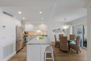 A modern kitchen with white cabinets, a stainless steel refrigerator, a granite countertop, and a dining area with wicker chairs and a table. - Gulf-view 2BR condo with balcony, pool, WiFi, and beach access. (Boca Grande)