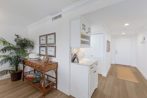 Een hal met een witte deur, een houten tafel met een plant erop, en ingelijste foto's aan de muur.