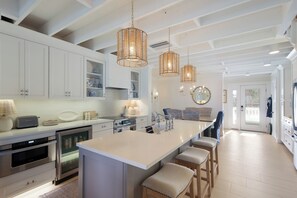 Cozinha moderna com armários brancos, ilha central com banquetas, iluminação pendente e vista para a sala de jantar.