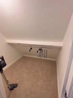 Une pièce avec un étagère fixée au plafond, sur laquelle sont rangés divers objets, un aspirateur posé sur le sol et un mur blanc.
