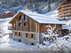 Eine Holzhütte mit Steinunterbau, zahlreichen Fenstern und einem mit Schnee bedeckten Dach, eingebettet in eine verschneite Landschaft mit weiteren Gebäuden.