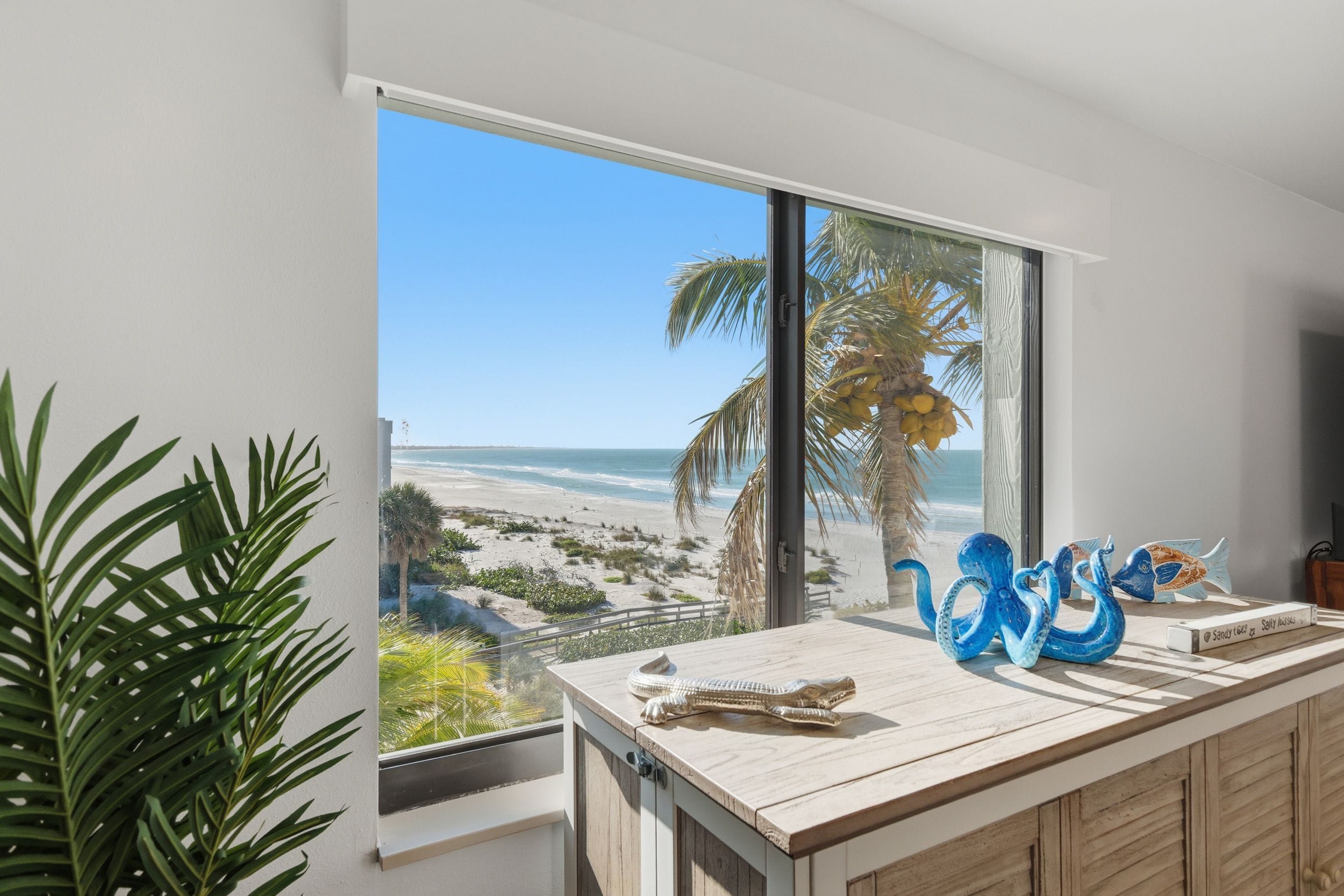 Una habitación con un ventanal grande con vista a una playa, una encimera de madera con adornos y una planta en maceta.