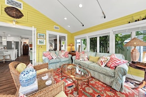 A living room with a sofa, coffee table, and wicker furniture. There is a kitchen visible in the background. - 271 Lee Cottage | Quaint Boca Grande Village Charm—Stroll to Shops, Dining & Island Life (Boca Grande)