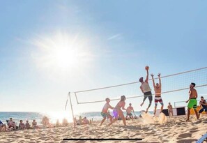 Ein Beachvolleyballspiel mit Spielern in Badesachen, einem Volleyballnetz und dem Meer im Hintergrund.