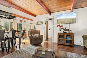 A spacious living room with a wooden ceiling, a dining area with chairs and a table, and a view of the outdoors through a window. - The Little Pine | Cozy 2BR | Crestline (Crestline)