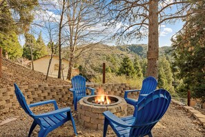 A fire pit with four blue chairs arranged around it, set against a backdrop of trees and a hillside. - The Little Pine | Cozy 2BR | Crestline (Crestline)