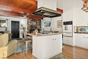 A cozy kitchen with a stone fireplace, wooden ceiling, and a small island with a sink. - The Little Pine | Cozy 2BR | Crestline (Crestline)
