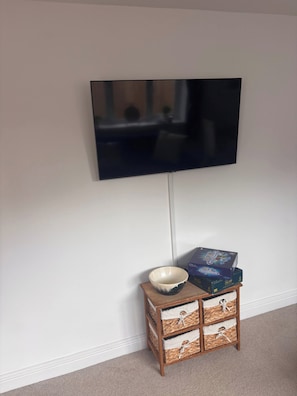 A television mounted on a wall, a small wooden side table with a bowl and a game box, and a carpeted floor. - Hidden Gem With Enclosed Garden and Parking (Sheringham)