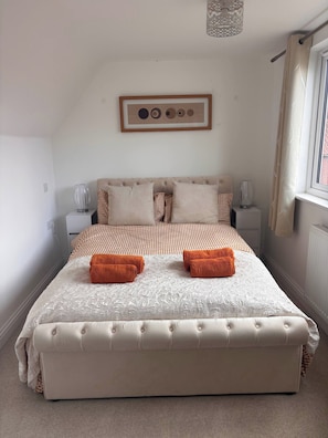 A neatly made bed with a checkered duvet, two bedside tables with lamps, a framed picture on the wall, and a window with curtains. - Hidden Gem With Enclosed Garden and Parking (Sheringham)