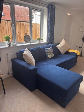 A blue sectional sofa with multiple cushions, a potted plant, a wall clock, and a window with curtains. - Hidden Gem With Enclosed Garden and Parking (Sheringham)
