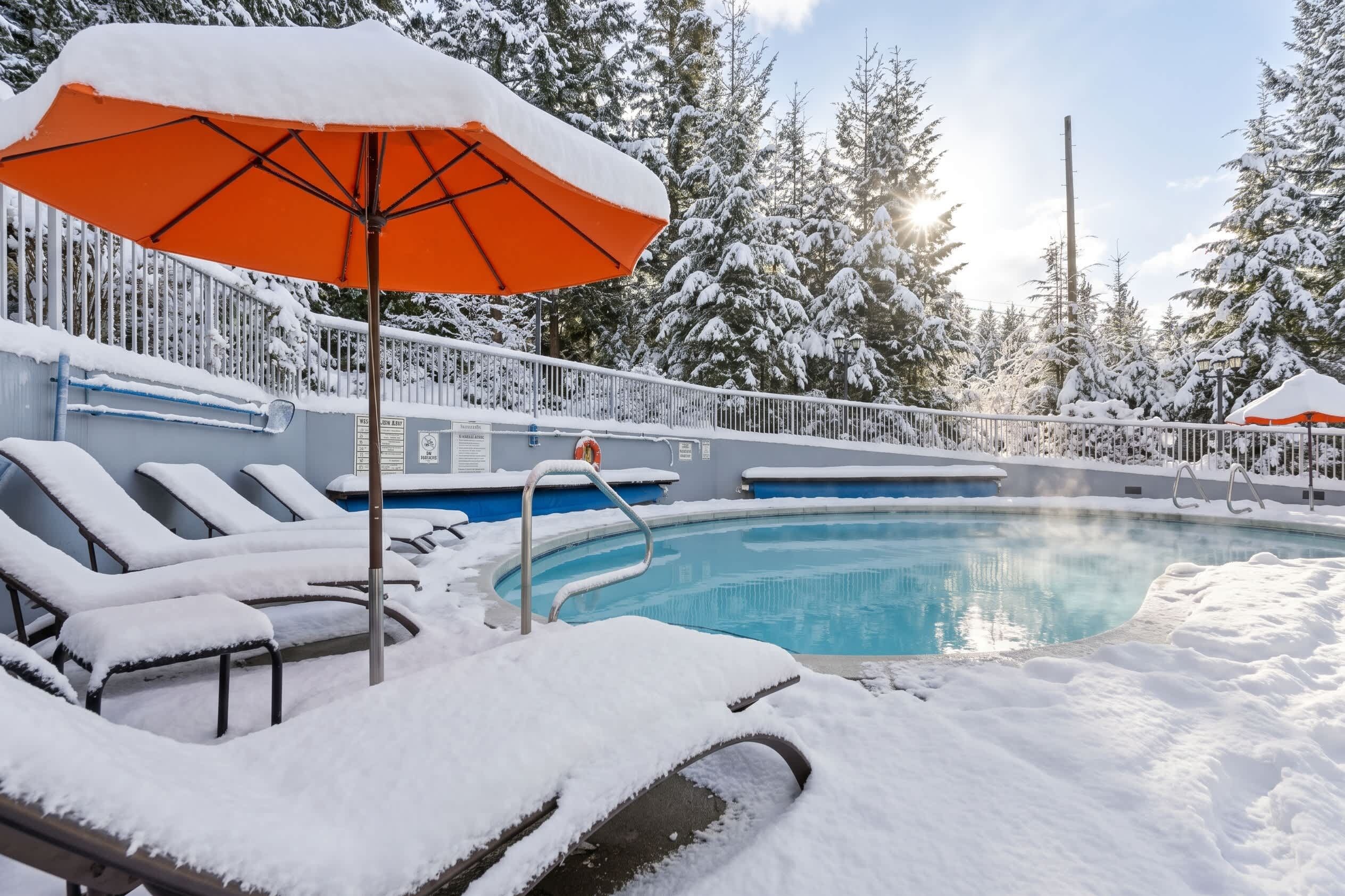 A snow-covered outdoor pool area with lounge chairs and an orange umbrella.